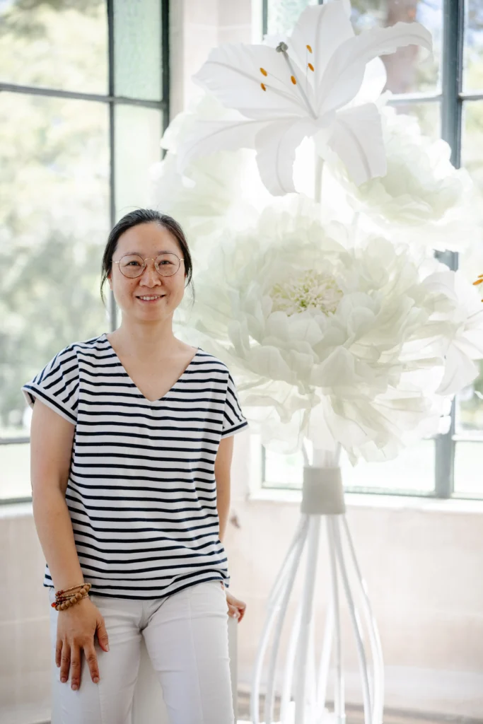 Artiste PaperBay devant une composition florale sculpturale en papier — lys et pivoine géants blancs — art floral événementiel sur mesure